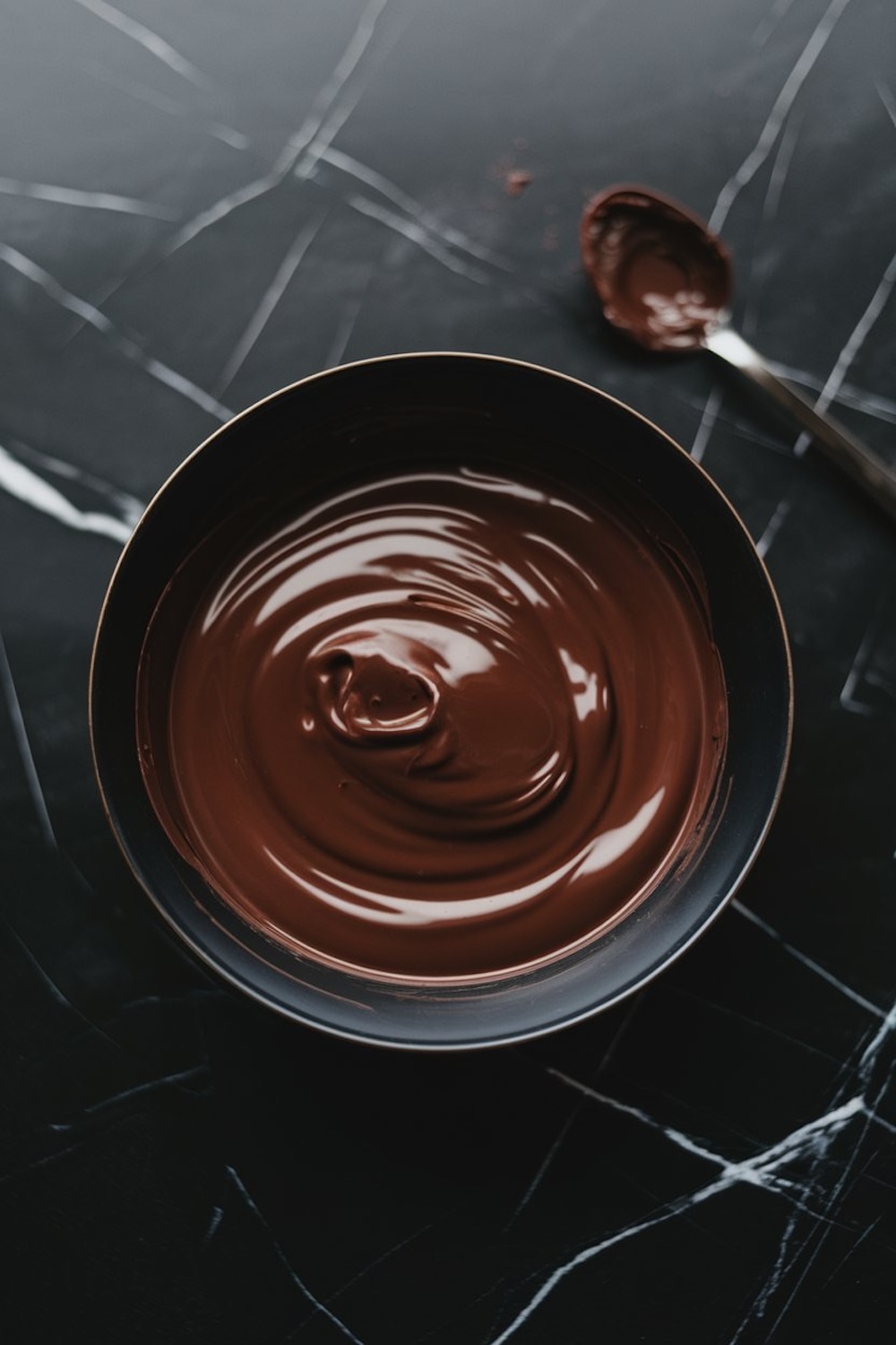 A bowl of smooth, melted dark chocolate ready for dipping, with a spoon resting nearby on a dark, marble countertop.