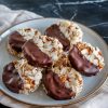 A plate of finished chocolate-dipped coconut cookies, highlighting their rich chocolate glaze, crispy almond slices, and coconut texture, presented on a beige napkin over a dark countertop.