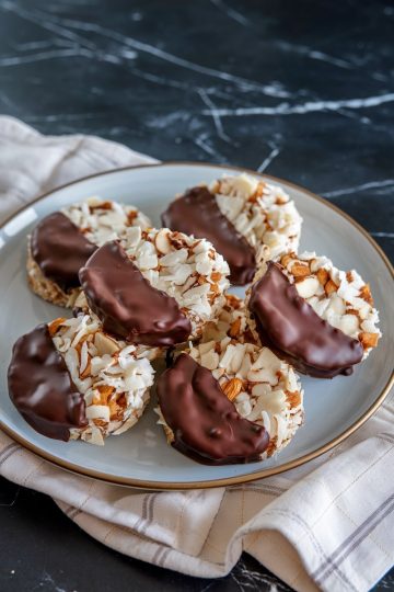 A plate of finished chocolate-dipped coconut cookies, highlighting their rich chocolate glaze, crispy almond slices, and coconut texture, presented on a beige napkin over a dark countertop.