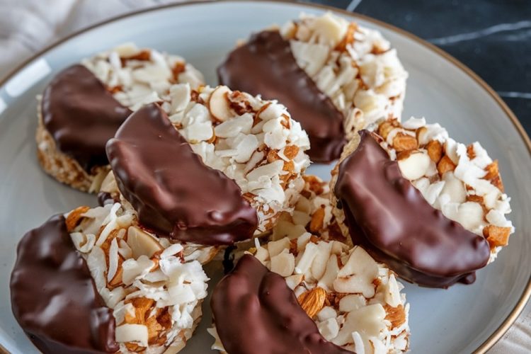 A plate of finished chocolate-dipped coconut cookies, highlighting their rich chocolate glaze, crispy almond slices, and coconut texture, presented on a beige napkin over a dark countertop.