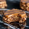 A close-up of a caramel pecan cluster on a glossy black marble surface. The treat features a base of pecans and chocolate, topped with a smooth caramel layer and finished with a chocolate drizzle. A bite has been taken, revealing the layers of chewy caramel and crunchy nuts.
