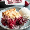 A slice of cherry dump cake served on a gray plate with vanilla ice cream. The dessert features a golden, flaky crust with vibrant, juicy cherry filling spilling onto the plate. A silver fork rests beside the cake. Background shows blurred cherries and a baking dish. Text overlay reads "Cherry Dump Cake."