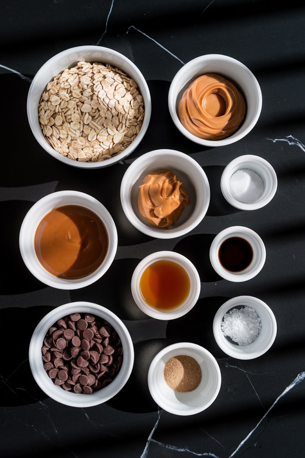 The image features neatly arranged ingredients in white bowls on a black marble surface. The ingredients include rolled oats, peanut butter, caramel sauce, chocolate chips, vanilla extract, salt, and other flavorings, emphasizing the simple components of the recipe.