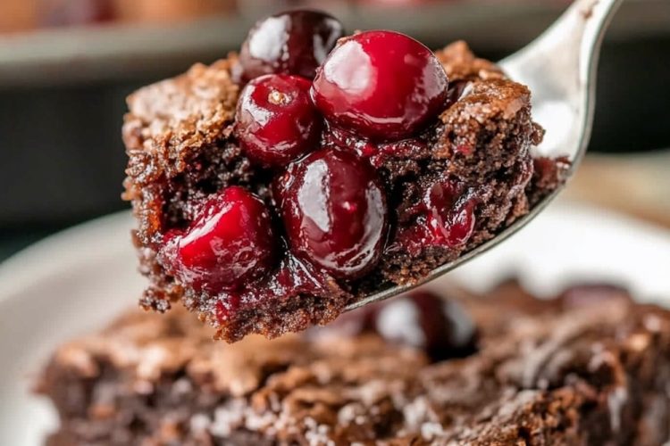 A close-up of a fork holding a bite of chocolate cherry dump cake, featuring a moist, fudgy texture filled with vibrant red cherries. The background shows a slice of the cake on a white plate, highlighting its rich chocolate layers and crackly crust.