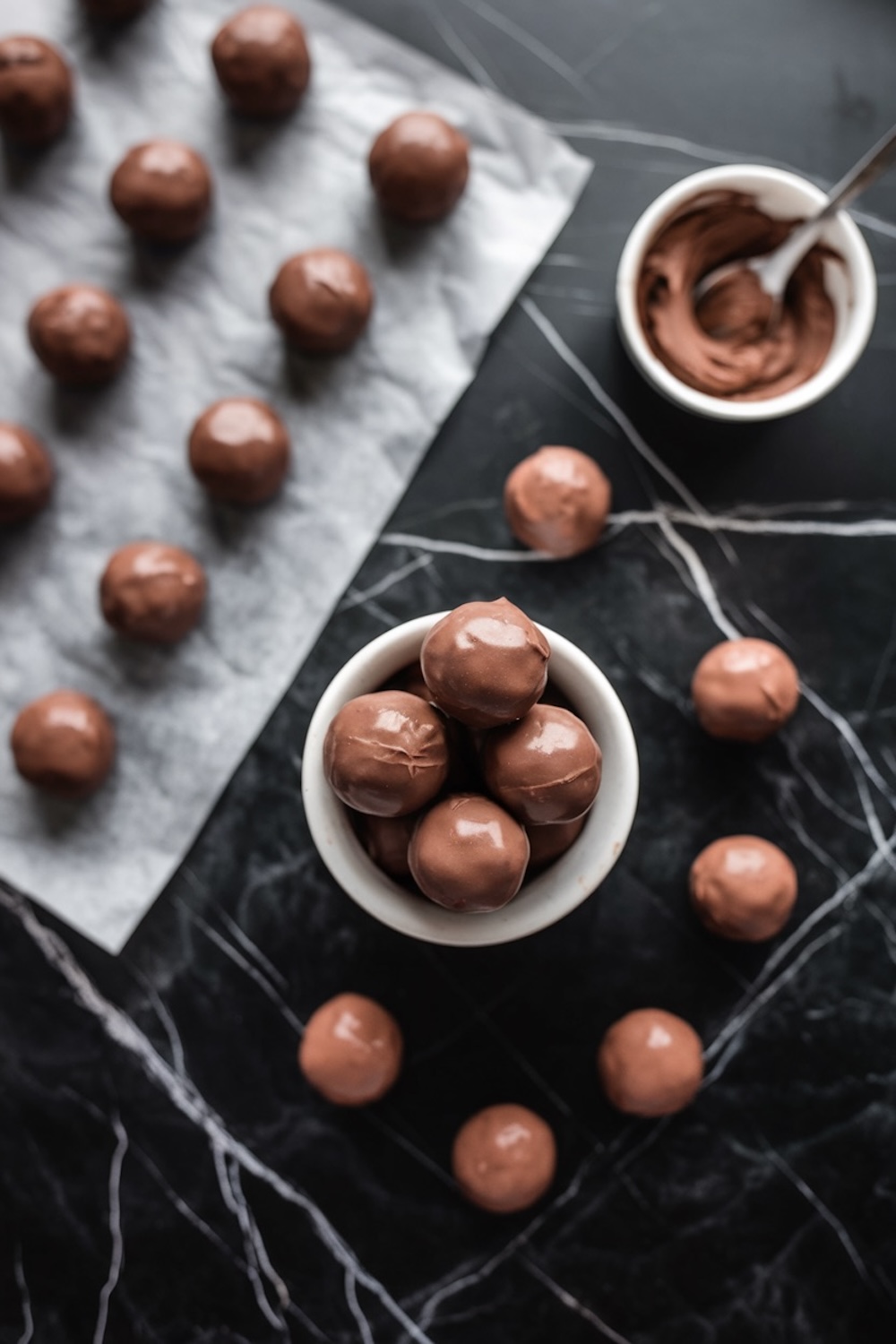 Rows of glossy chocolate-coated balls resting on parchment paper, with a small white bowl filled with more of these chocolate treats. The scene is set on a dark marble surface with elegant contrast.