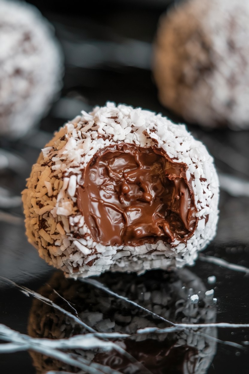 A close-up of a single chocolate coconut no-bake ball, bitten to showcase the gooey chocolate center. The shredded coconut exterior creates a striking contrast against the dark background.