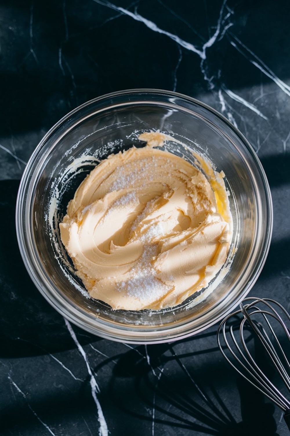 A creamy mixture of butter and sugar in a glass bowl, ready for baking, with sunlight streaming over a black marble surface.