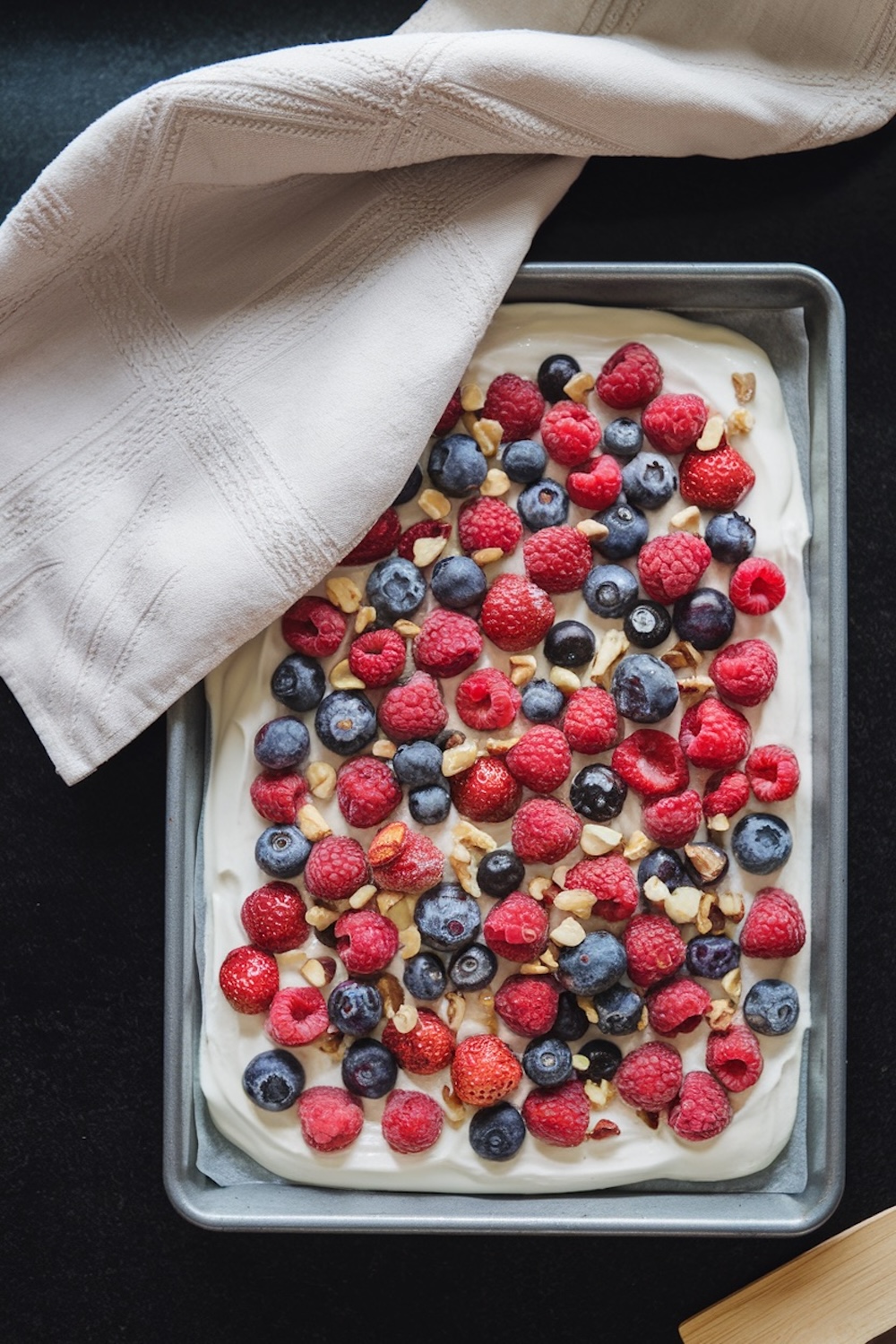 A baking pan filled with a layer of creamy yogurt, topped with fresh raspberries, blueberries, strawberries, and chopped nuts. A white textured kitchen towel partially drapes over the corner of the pan, placed on a dark surface.