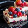 Close-up of frozen yogurt bark topped with fresh strawberries, blueberries, raspberries, and chopped nuts on a black marble surface. The yogurt features vibrant berry pieces and crunchy toppings.