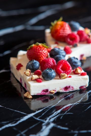 Close-up of frozen yogurt bark topped with fresh strawberries, blueberries, raspberries, and chopped nuts on a black marble surface. The yogurt features vibrant berry pieces and crunchy toppings.
