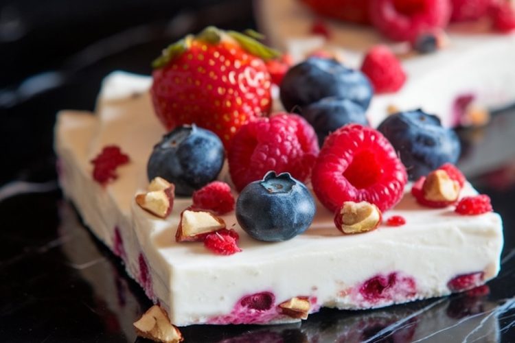 Close-up of frozen yogurt bark topped with fresh strawberries, blueberries, raspberries, and chopped nuts on a black marble surface. The yogurt features vibrant berry pieces and crunchy toppings.
