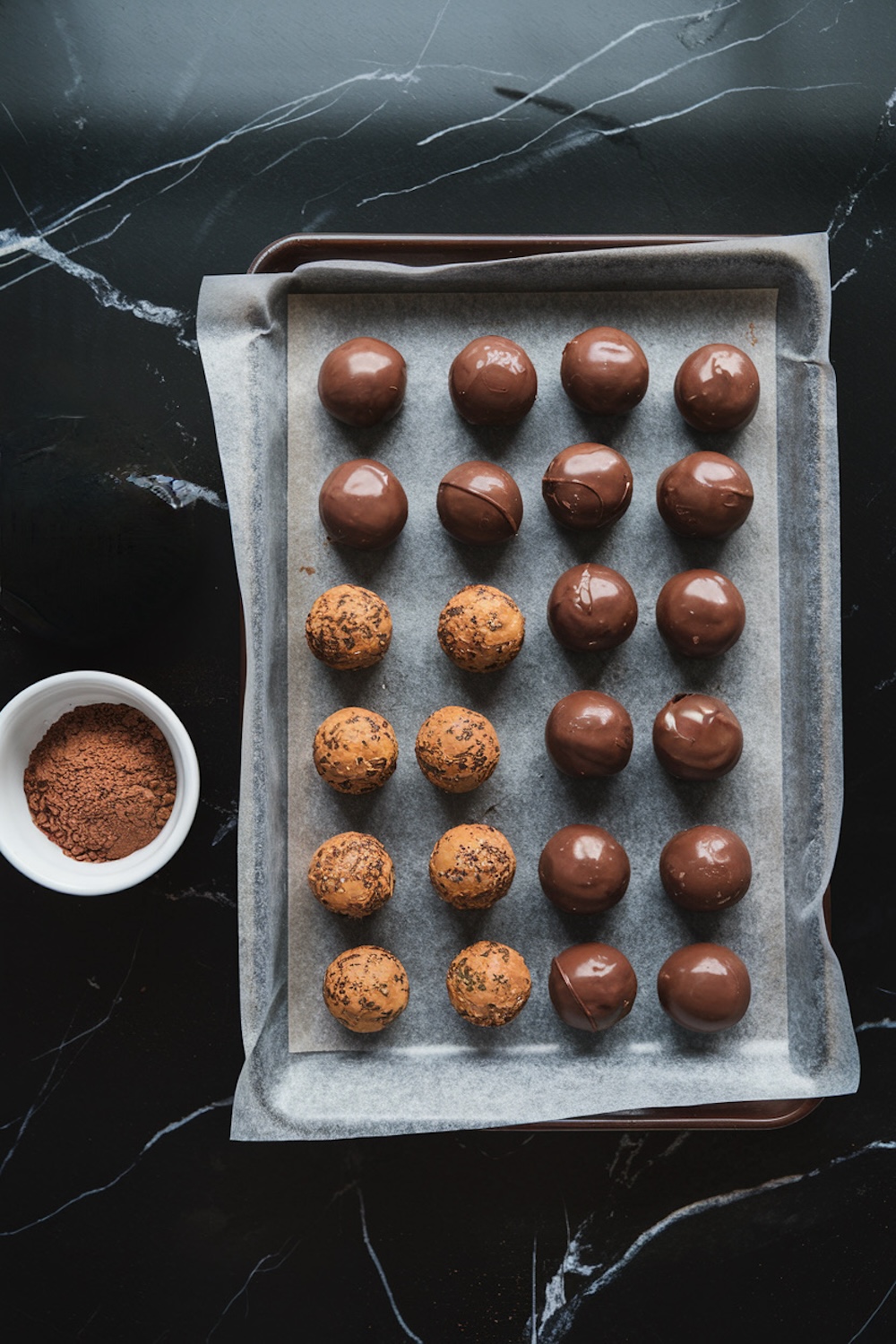 Tray of chocolate truffles in two stages on parchment paper, placed on a black baking sheet. Half are rolled in cocoa powder, while the other half are coated in glossy chocolate. A small white bowl of cocoa powder sits nearby.