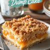 A close-up of a slice of papaya cream dump cake on a beige plate, featuring layers of creamy filling, fresh papaya chunks, and a golden crumbly topping. A silver fork rests beside the slice, and the background includes blurred details of a white mug and additional dessert plates.