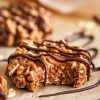 A single image showing a peanut butter chocolate caramel cookie with a bite taken, revealing a gooey caramel center, topped with chocolate drizzle, and scattered oats on parchment paper.