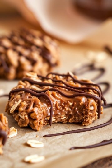A single image showing a peanut butter chocolate caramel cookie with a bite taken, revealing a gooey caramel center, topped with chocolate drizzle, and scattered oats on parchment paper.