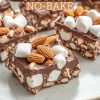 Close-up of no-bake rocky road clusters on a beige plate, featuring a thick chocolate base topped with marshmallows and whole almonds, showcasing a rich, crunchy texture.