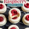 Close-up of white chocolate raspberry thumbprint cookies arranged on a black marble surface. The cookies are topped with fresh raspberries and coated in smooth white chocolate. One cookie has a bite taken out, revealing a soft, buttery interior.