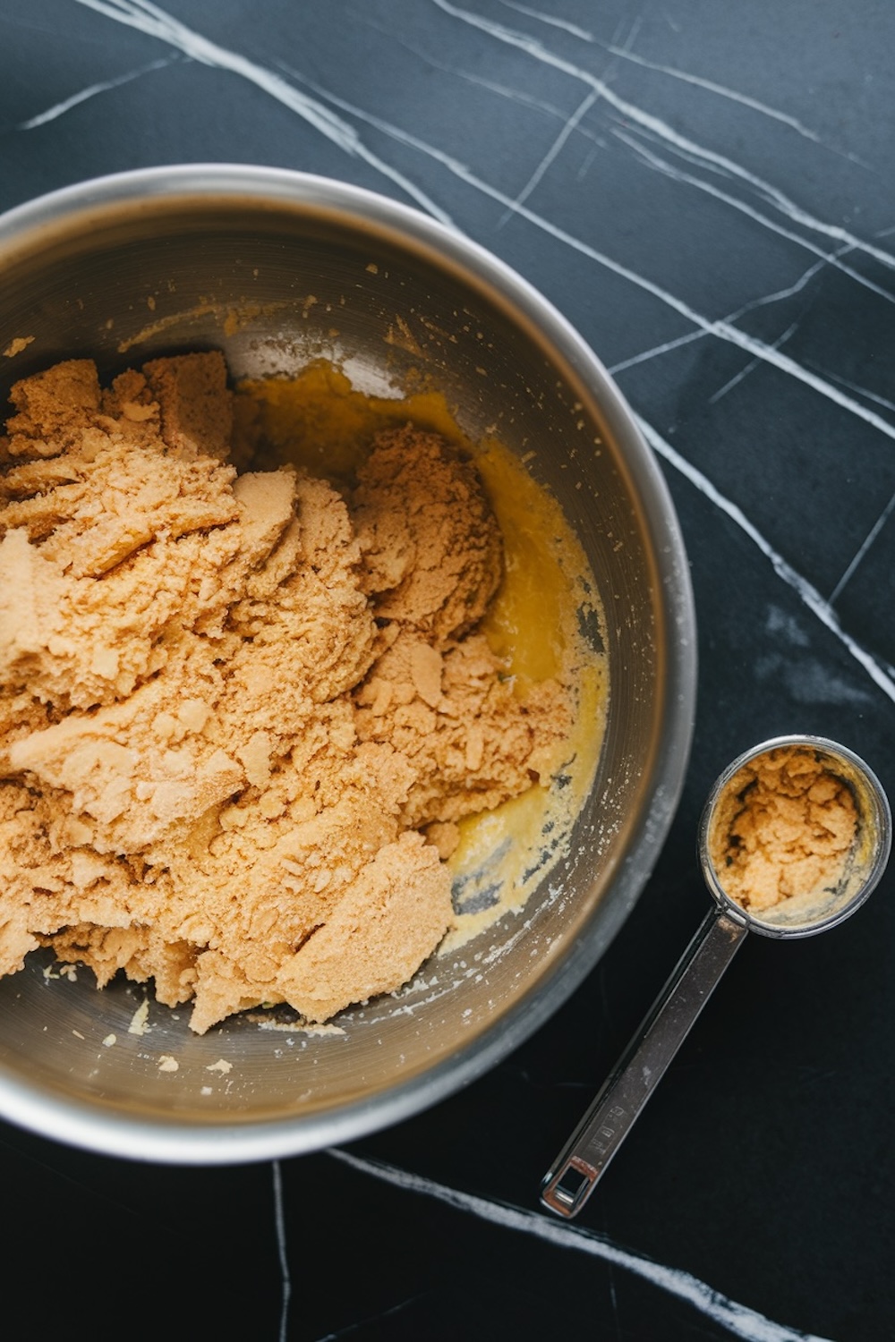 A mixing bowl with a crumbly cookie mixture for white chocolate raspberry cookies. A metal measuring spoon sits beside the bowl on a black marble surface.
