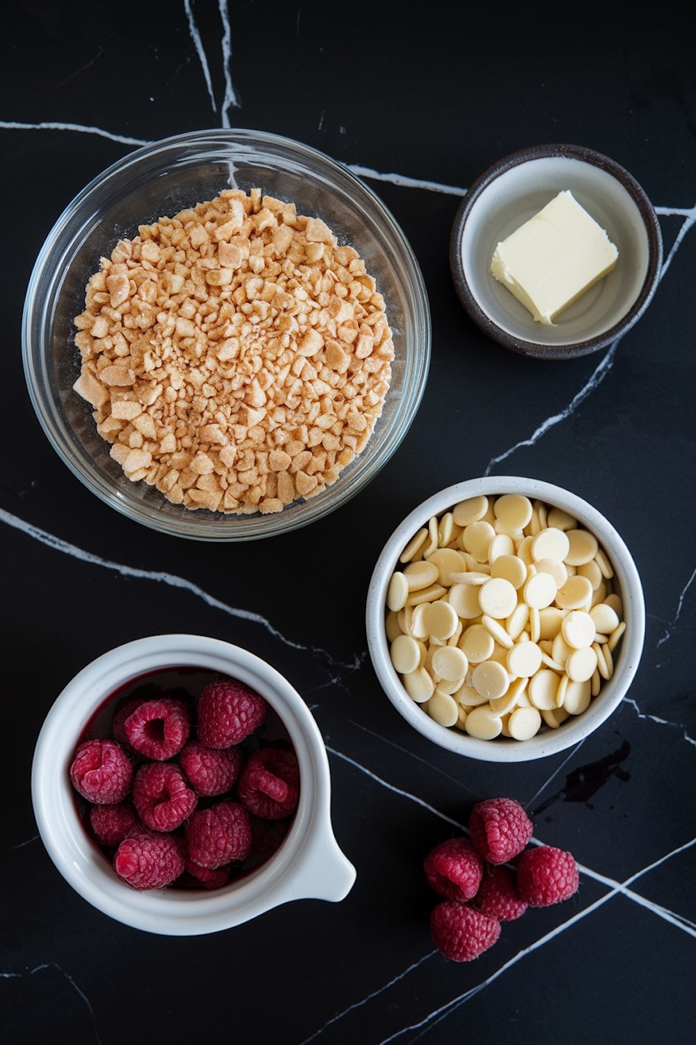 Flat-lay view of ingredients for white chocolate raspberry thumbprint cookies. A bowl of crushed cookie crumbs, white chocolate chips, fresh raspberries, and a small portion of butter are displayed on a black marble countertop.