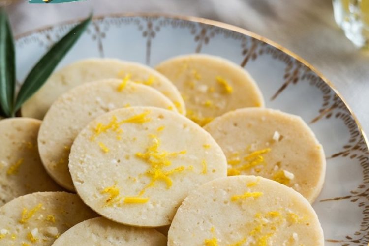 A close-up of "Zesty Cashew Lemon Treats," showcasing golden round cookies on a decorative porcelain plate, sprinkled with fresh lemon zest. The background includes soft lighting and blurred glasses of refreshing beverages.