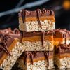 Stacked chocolate caramel Rice Krispie treats on a cooling rack, featuring glossy caramel and chocolate layers dripping slightly. Crisp rice cereal base adds contrast to the rich toppings. Keywords: stacked Rice Krispie treats, caramel chocolate layers, cooling rack, dessert close-up.