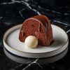 A single slice of chocolate Bundt cake drizzled with glossy chocolate glaze, served with a scoop of vanilla ice cream on a white plate with a gold rim. The plate sits on a sleek black marble surface, creating a contrast that highlights the dessert’s rich, moist texture and indulgent presentation.