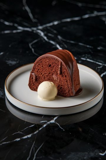 A single slice of chocolate Bundt cake drizzled with glossy chocolate glaze, served with a scoop of vanilla ice cream on a white plate with a gold rim. The plate sits on a sleek black marble surface, creating a contrast that highlights the dessert’s rich, moist texture and indulgent presentation.
