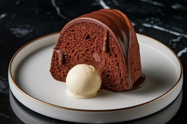 A single slice of chocolate Bundt cake drizzled with glossy chocolate glaze, served with a scoop of vanilla ice cream on a white plate with a gold rim. The plate sits on a sleek black marble surface, creating a contrast that highlights the dessert’s rich, moist texture and indulgent presentation.