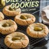 Freshly baked dill pickle cookies cooling on a wire rack. The cookies have a soft, golden exterior with slight cracks and are topped with green pickle slices. In the background, a jar of pickles and a folded kitchen towel complete the scene.
