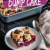 A plated slice of triple berry dump cake with a crumbly, golden topping and berry filling, placed on a white plate with a blue rim. The background features a baking pan of the cake and a beige napkin with a silver spoon on a black marble countertop. Overlay text reads “Triple Berry Dump Cake.”