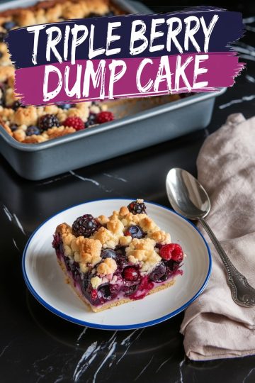 A plated slice of triple berry dump cake with a crumbly, golden topping and berry filling, placed on a white plate with a blue rim. The background features a baking pan of the cake and a beige napkin with a silver spoon on a black marble countertop. Overlay text reads “Triple Berry Dump Cake.”