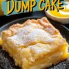 A close-up of a single slice of lemon dump cake on a black plate, dusted with powdered sugar. The vibrant yellow filling contrasts with the golden crust. Overlay text reads: “Lemon Dump Cake.” Fresh lemons and lemon sauce appear blurred in the background.