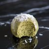 Close-up of a single matcha coconut snack ball with a bite taken out, revealing its fluffy, green interior and topped with a sprinkle of shredded coconut for a decorative finish.