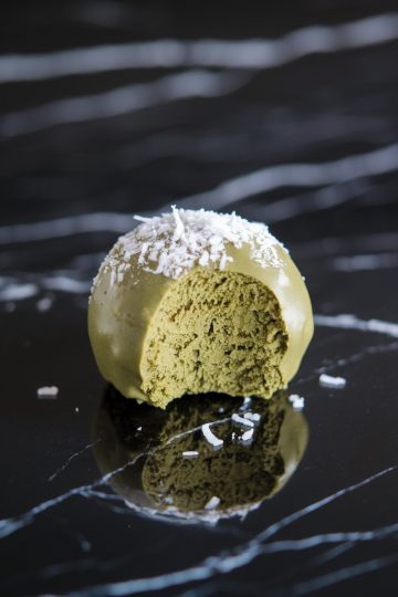 Close-up of a single matcha coconut snack ball with a bite taken out, revealing its fluffy, green interior and topped with a sprinkle of shredded coconut for a decorative finish.