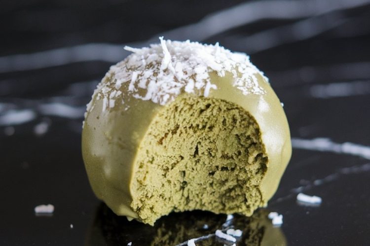 Close-up of a single matcha coconut snack ball with a bite taken out, revealing its fluffy, green interior and topped with a sprinkle of shredded coconut for a decorative finish.