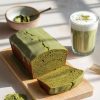 A beautifully styled matcha pound cake on a wooden board, sliced to reveal its vibrant green interior and topped with a smooth matcha glaze. The setup includes a steaming matcha latte in a glass and a bowl of matcha powder in the background, with sunlight casting soft shadows, creating a warm and inviting scene perfect for highlighting the cake's earthy flavors and aesthetic appeal.