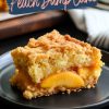 A close-up of a single slice of peach dump cake served on a black plate. The warm peaches, syrupy filling, and crispy buttery topping create a visually appealing dessert perfect for casual or holiday servings.