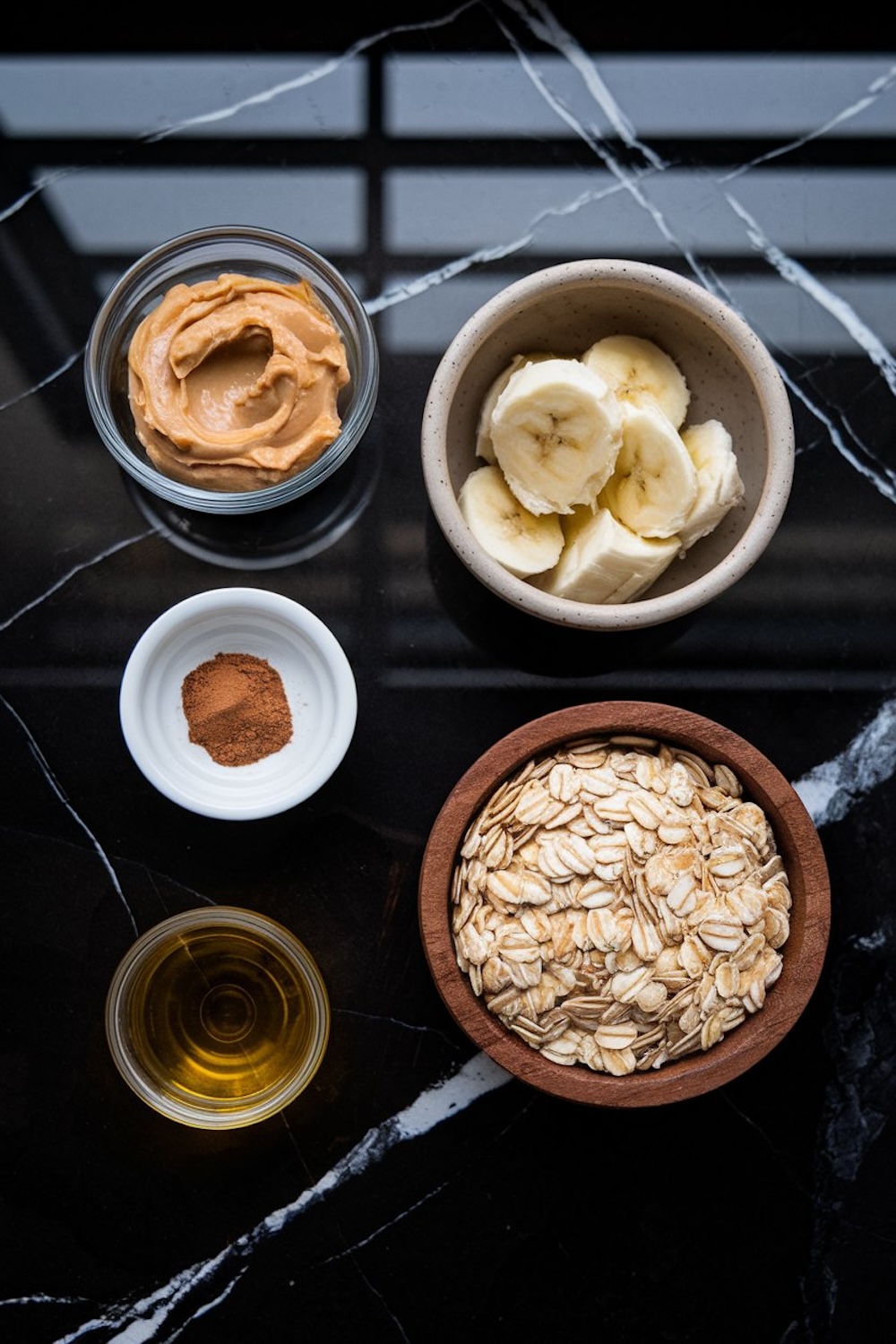 A flat lay of ingredients for energy balls on a black marble surface: creamy peanut butter in a glass bowl, banana slices in a ceramic dish, a small bowl of cinnamon, a wooden bowl of rolled oats, and a cup of golden syrup or honey.