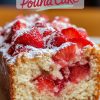 Close-up of a strawberry pound cake slice with fresh strawberries embedded in the soft, fluffy cake, topped with powdered sugar. The cake has juicy red strawberries throughout, creating a vibrant and moist texture. Overlay text reads “Strawberry Pound Cake.”