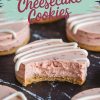 A close-up of a single strawberry cheesecake cookie with a bite taken out, revealing the creamy pink filling and crumbly cookie crust. The icing drizzle and visible strawberry pieces enhance the dessert’s visual appeal, making it an irresistible treat.