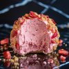 A detailed view of a bitten strawberry shortcake ball, showing the smooth, whipped pink filling surrounded by a crunchy crumb coating with scattered strawberry pieces.