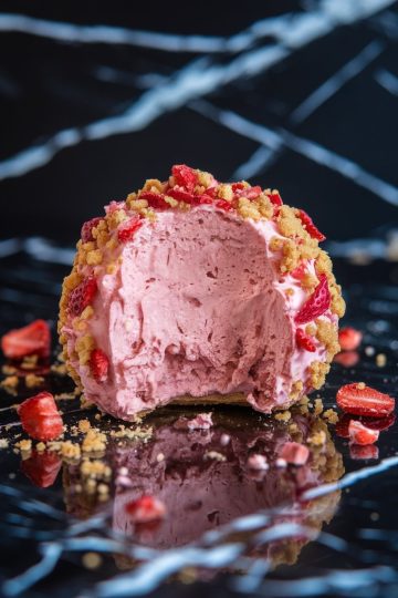 A detailed view of a bitten strawberry shortcake ball, showing the smooth, whipped pink filling surrounded by a crunchy crumb coating with scattered strawberry pieces.
