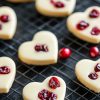 A close-up view of multiple white chocolate cranberry hearts cooling on a wire rack, with vibrant red cranberries scattered around for contrast.