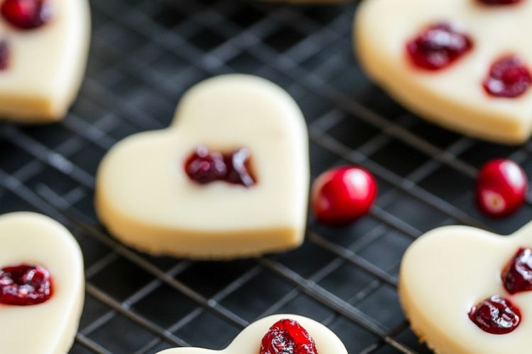 A close-up view of multiple white chocolate cranberry hearts cooling on a wire rack, with vibrant red cranberries scattered around for contrast.