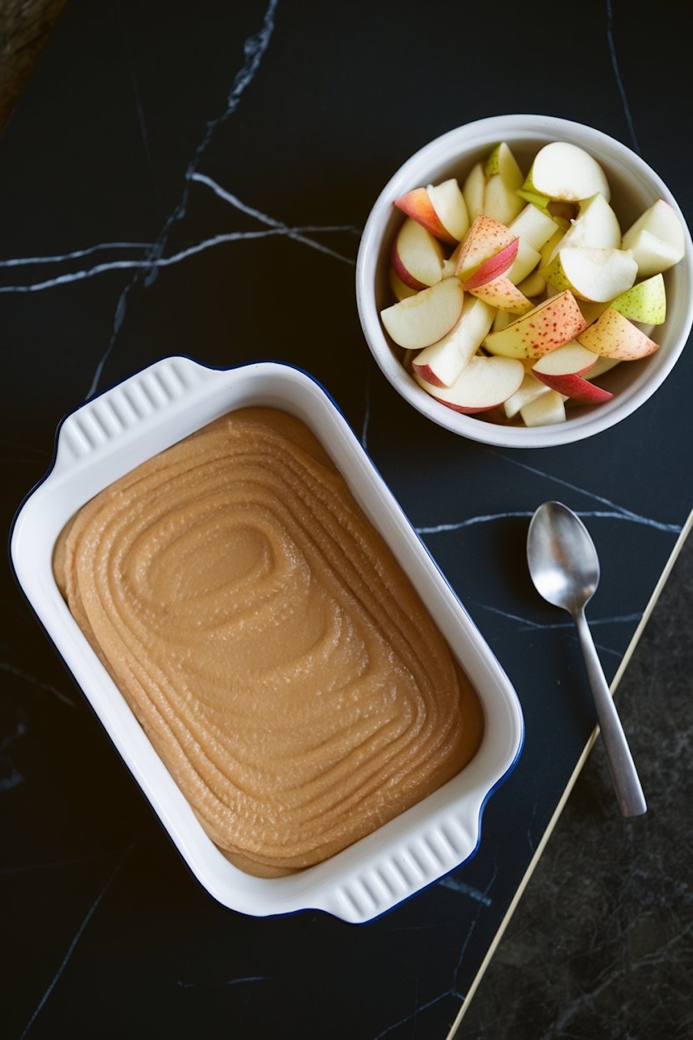 A top-down view of a baking setup, showcasing a rectangular white baking dish filled with smooth, swirled apple puree. Next to it is a bowl of fresh apple and pear slices, with a silver spoon resting on the black marble surface.