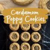 A batch of freshly baked cardamom poppy seed cookies cooling on a black wire rack, arranged neatly on a black marble surface. The cookies have a golden-brown color with a sprinkle of poppy seeds in the center.