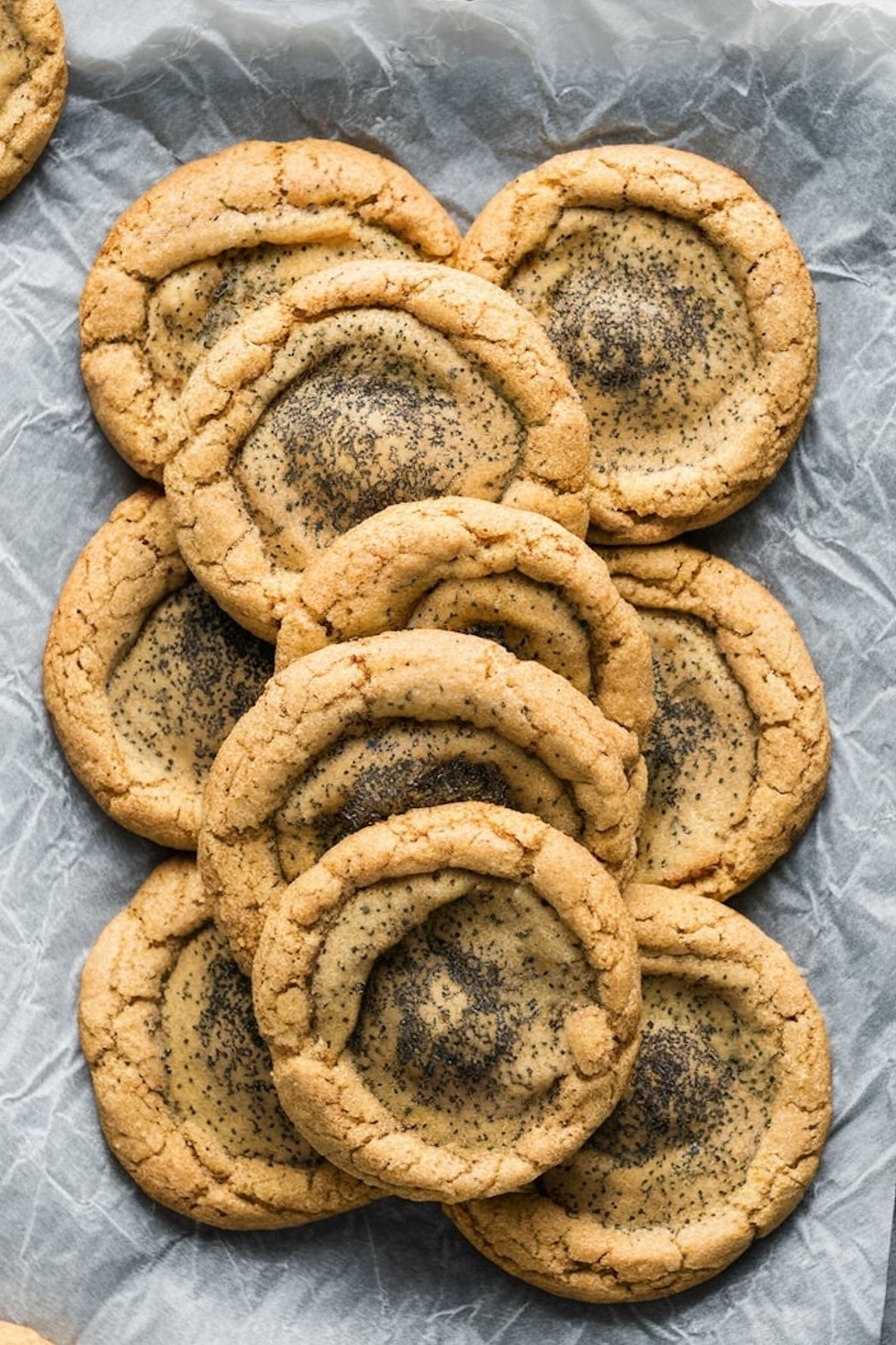  A close-up view of golden cardamom poppy seed cookies arranged in a neat stack on crinkled parchment paper, highlighting their crisp edges and textured centers.