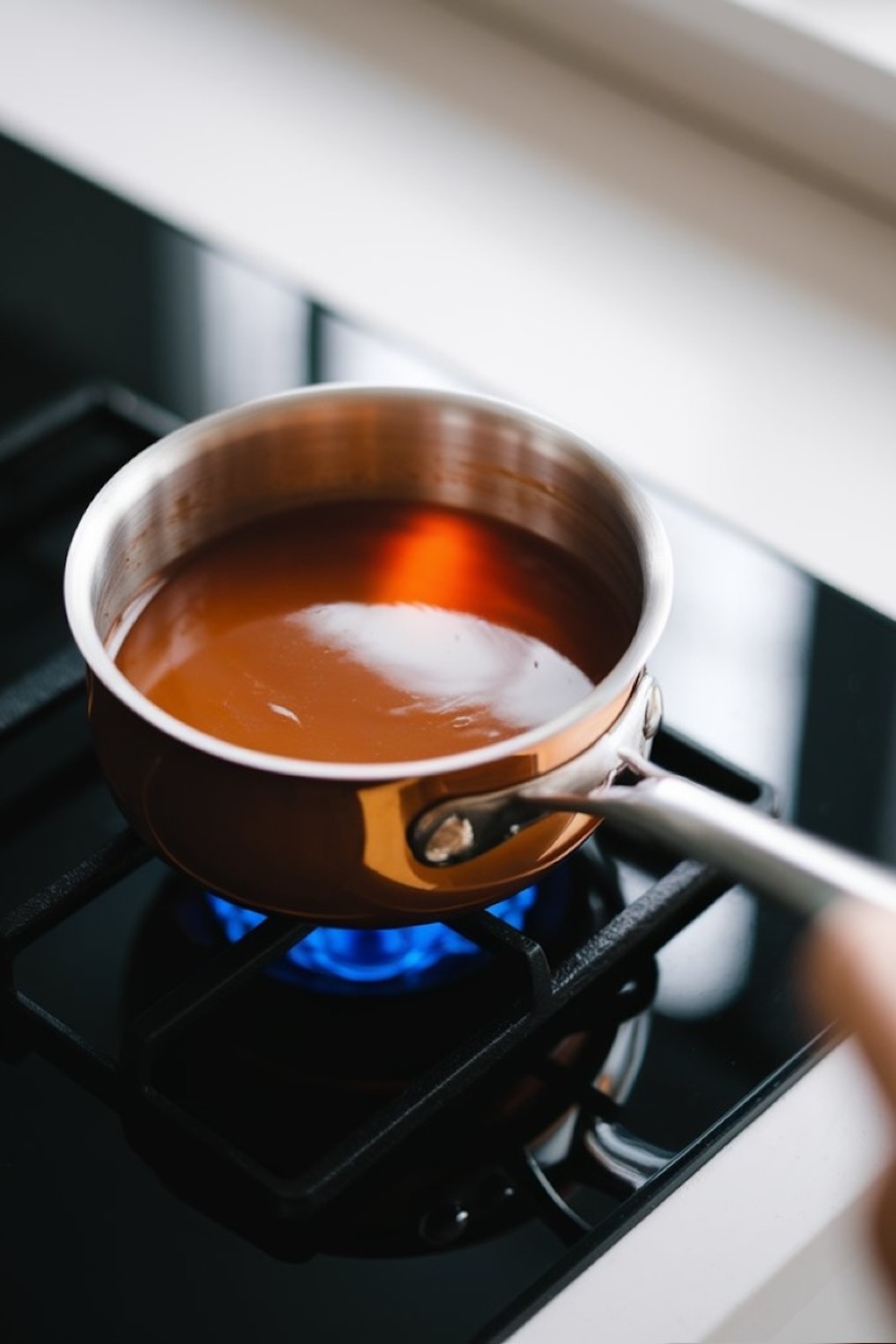 A small saucepan of melted caramel sauce heating on a gas stove, with warm light reflecting off the shiny surface of the pan.