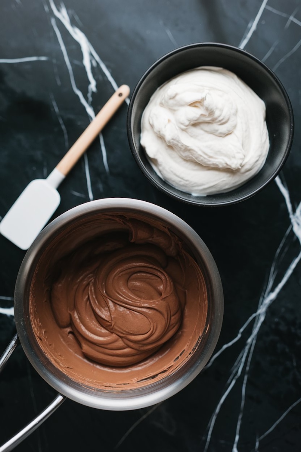A preparation scene showing a bowl of whipped cream and a saucepan filled with smooth, glossy chocolate mousse. A white silicone spatula rests nearby on the dark marble surface, highlighting the steps to create the cake’s mousse filling.