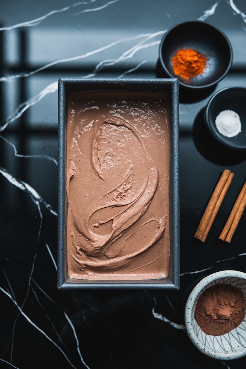 A loaf pan filled with chocolate chipotle ice cream, featuring a creamy, swirled texture. The pan is placed on a black marble surface with small bowls of spices and cinnamon sticks arranged beside it.